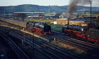 Two substantial red and black, East German steam locomotives pass on a multi-track railway, with lineside industrial buildings behind and tree-covered hills in the distance.